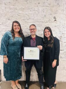 A man holding a certificate award is wearing a navy jacket and is standing against a  white brick wall. There is a woman in business formal outfits on either side of the man. 