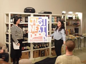 Two brunette women stand in front of a shelf of red wine bottles. In between them is an easel with a large rendering of the Just Next Door logo