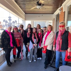 A group of 11 woman of all ages wearing valentines day pink scarves and glasses stand on a front porch.